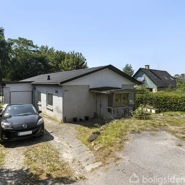 En sort bil holder parkeret foran et hvidt hus med fladt tag. Huset har en garage til venstre, og omgivelserne består af buske og træer.