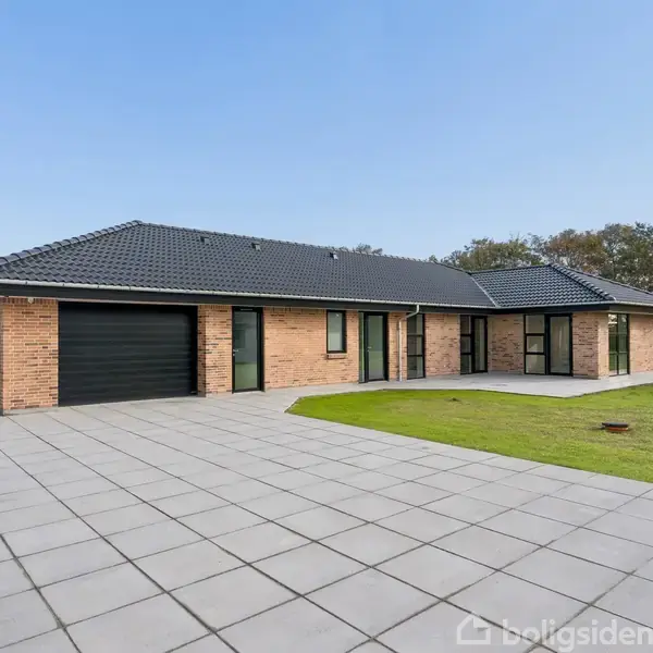 En moderne murstensvilla står på en flisebelagt indkørsel. Villaen har et sort tag og integreret garage. Den grønne græsplæne omgiver huset, og træer ses i baggrunden.