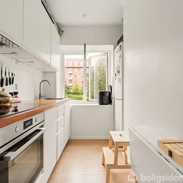 Et smalt, moderne køkken har hvide skabe, en indbygget ovn og kogeplade. Køkkenet er lyst og har et vindue med udsigt til grønne træer og en bygning.