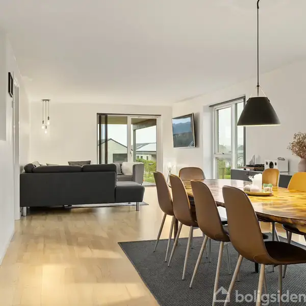 En moderne spisestue med et stort ovalt træbord omgivet af otte stole, på en grå tæppe. I baggrunden ses en stue med en sofa, fjernsyn, og store vinduer med udsigt.