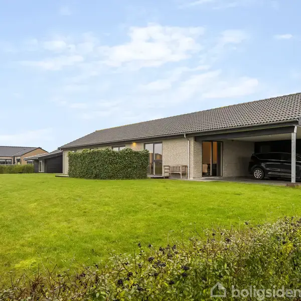 Hus med enkelt-etage står stille i en grøn have, omgivet af en lav hæk. Til højre er en carport med en bil parkeret. Himmel er let overskyet.
