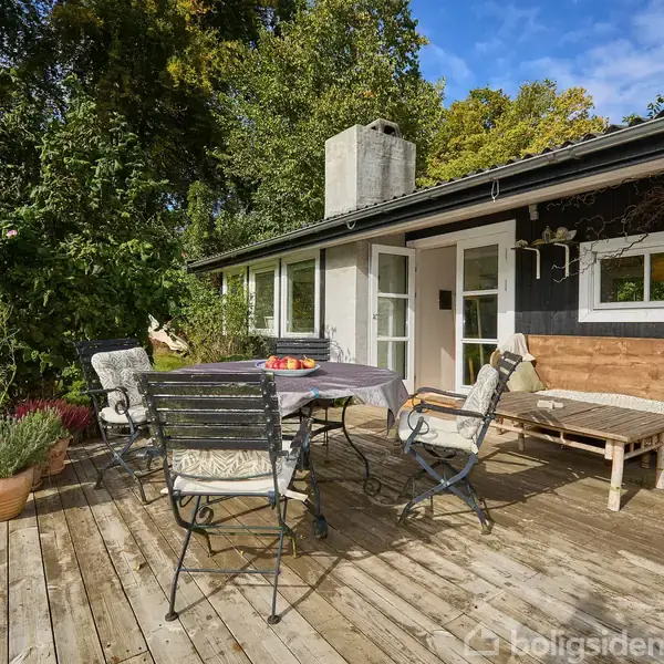 Terrasse med bord og stole står på trædæk foran et sort-hvidt hus. En sofa med puder er ved siden af bordet. Omgivet af grønne buske og træer.