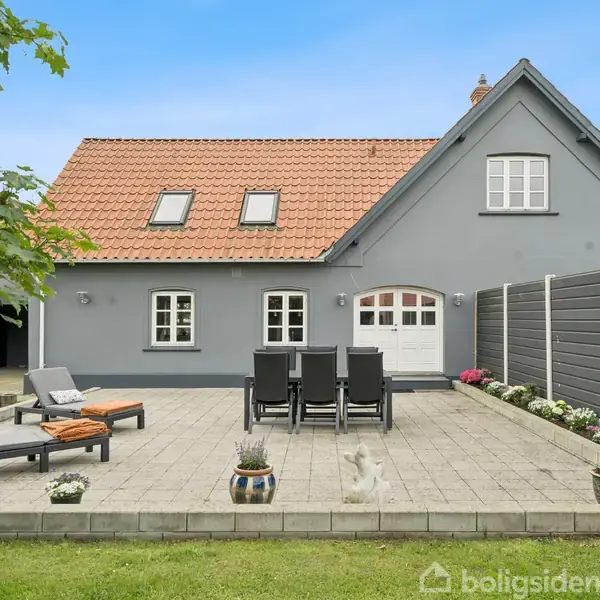 Gråmalet hus med rødt tegltag, omkranset af en terrasse med havestole og liggestole. Haven er grøn og omkranset af et hegn, med blomsterpotter pyntende på terrassen.