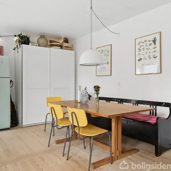Et træbord med to gule stole og en sort bænk står i et lyst køkken. I baggrunden ses et retro køleskab, skab og vægkunst. En hængelampe belyser spiseområdet.