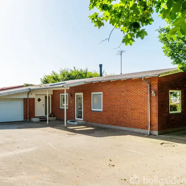 Rødt murstenshus med fladt tag, stor garage til venstre. Huset står på et åbent asfaltområde omgivet af grønne træer og buske. Sollys skaber en lys, indbydende atmosfære.