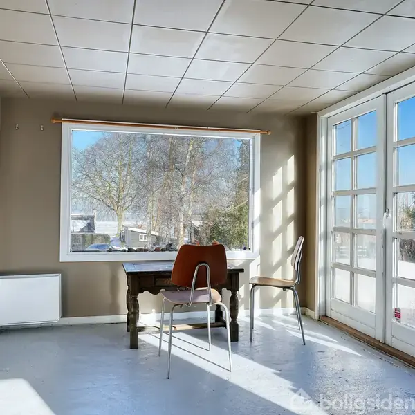 Et lyst rum med et træbord og to stole ved siden af store vinduer. Udenfor ses træer og huse i vinterlandskab. En radiator står ved væggen.