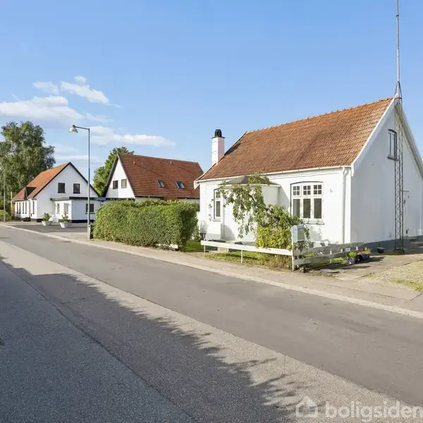 Et rækkehus med hvid facade og rødt tag står langs en stille vej. Til venstre ses en række lignende huse og grønne hække under en klar blå himmel.