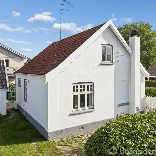 Hvidt hus med rødt tegltag står i stille boligområde omgivet af grønne hække og græsplæner. Husets vinduer er traditionelle med sprosser, og der ses en høj skorsten.