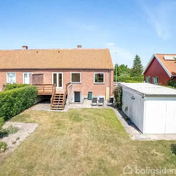 Rødstenshus med tag i teglsten og høje vinduer, træterrasse og trappe, beliggende i en velplejet have med garage til højre, omgivet af grønne hække og nabohuse.