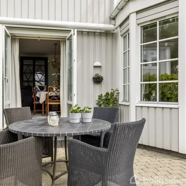 Rundt havebord omgivet af fire fletstole står på en flisebelagt terrasse foran en smuk gråmalet træbygning. Glasdøre åbner ind til et indendørs spiseområde med lysekrone.