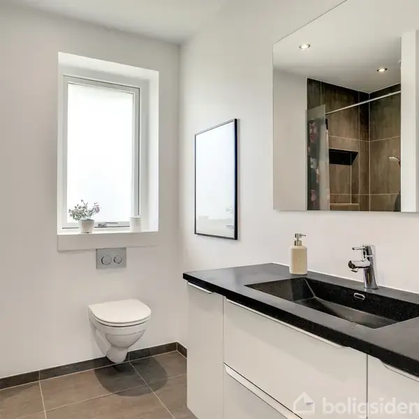 Toilet hænger på væggen, stille i et moderne badeværelse med brune fliser. Ved siden af er en vask med sort bordplade og spejl. Vindue i rummet. Ingen tekst set.