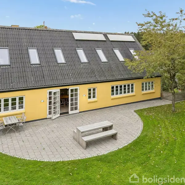 En gul, to-etagers bolig med vinduer i taget, har en stor, flisebelagt terrasse. Der står et havebord og bænk på terrassen, omringet af en velplejet græsplæne og træer.