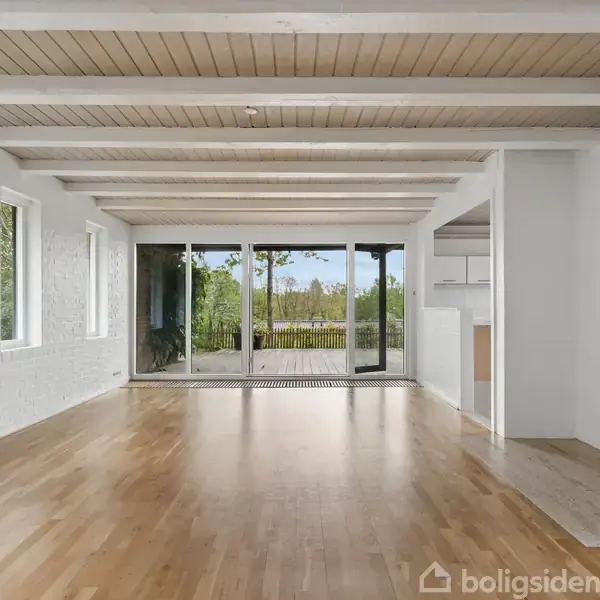 Stue med trægulv og loftsbjælker, store vinduer og glasdør, uden møbler. Pejs indbygget i væggen til højre. Udsigt til en terrasse og grønne omgivelser udenfor.