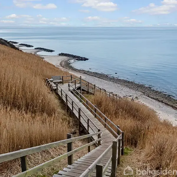 Trægangbro fører ned til stenstrand omgivet af højt græs. Havet strækker sig ud mod horisonten med let skyet himmel.