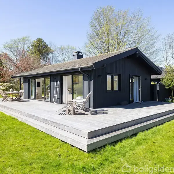 Et moderne sommerhus med store vinduer står på en træterrasse omgivet af en grøn græsplæne og træer. Der er havemøbler på terrassen i en solrig have.