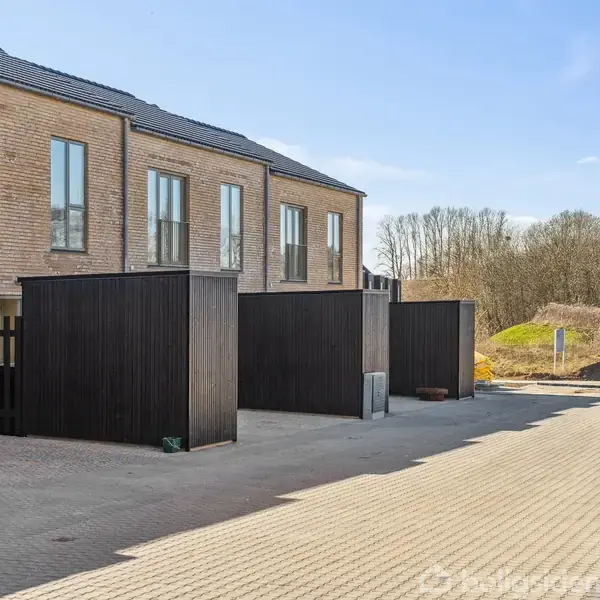 Rækkehus bygget af mursten; står i stille boligområde med parkeringsplads foran. En person i orange arbejdstøj går i baggrunden nær et grønt område og træer.