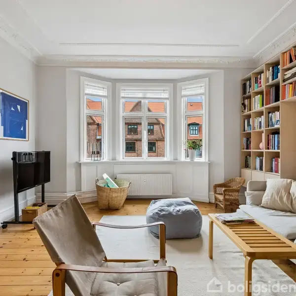 Stue med lyse vægge; sofa og lænestole arrangeret omkring et sofabord. Højre væg har en bogreol. Store vinduer med udsigt til røde taghuse. Kurve med puder på gulvet.