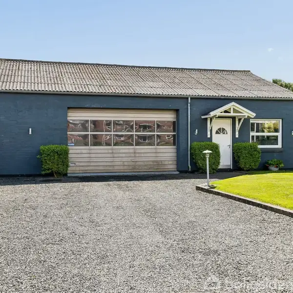 Et enfamiliehus med blå facade og gråt tag ses forfra. Huset har en garage med en stor port til venstre og en hvid hoveddør med et lille halvtag. Foran huset er der en grusbelagt indkørsel og en velplejet græsplæne.