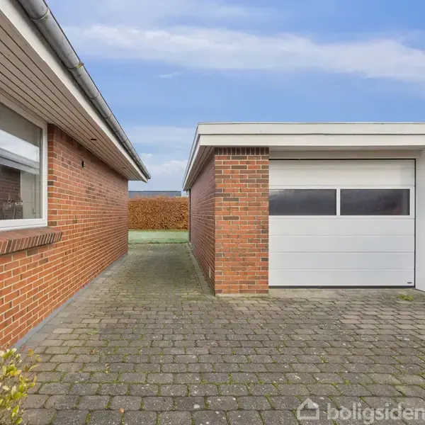 Garagebygning med hvid garageport står på flisebelagt indkørsel. Til venstre ses husvæg med vinduer; til højre og bagved er der hække. Himlen er blå med nogle skyer.