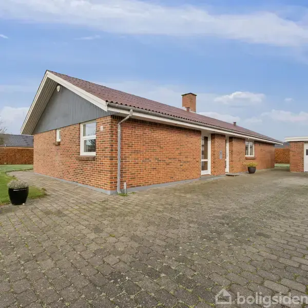 Et rødt murstenshus med tagsten står på en flisebelagt gårdsplads. Der er to sorte blomsterkrukker til venstre. En enkelt garage ses til højre i billedet.