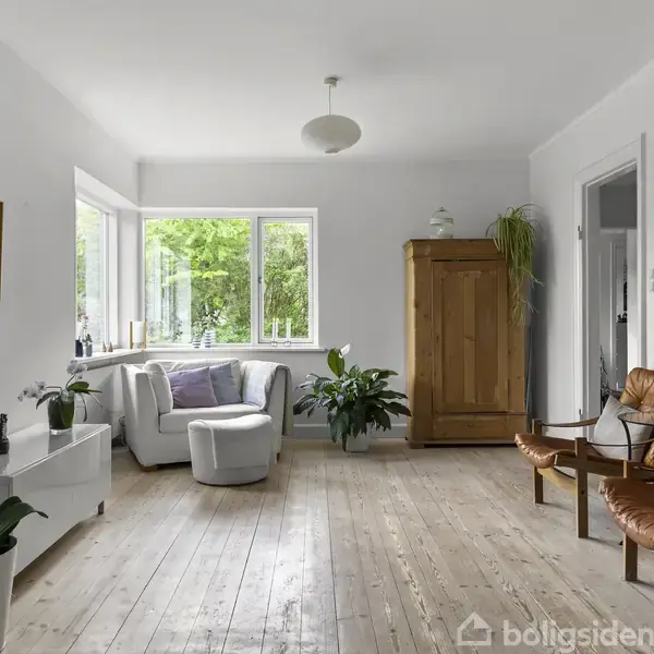 Et lyst rum indeholder en hvid lænestol ved vinduet, planter, en hvid kommode og et træskab. To brune lænestole står ved væggen med minimalistisk kunst.