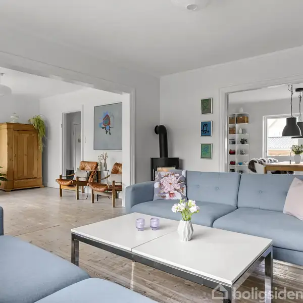 En stue med blå sofaer arrangeret omkring et hvidt sofabord; en vase med blomster står midt på bordet. I baggrunden ses stole, brændeovn og et skab. Ingen tekst.