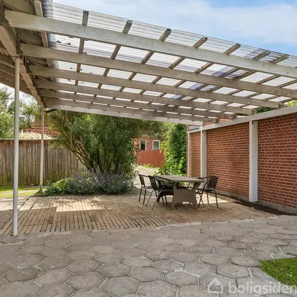 Terrassemøbler står under en overdækket udendørs terrasse i en have. Terrassen har røde murstensvægge, flisegulv og et gennemsigtigt tag. Bagerst ses grønne buske og et hegn.