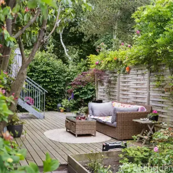 Rattanmøbler står på en træterrasse med puder. Terrassen er omgivet af frodige planter og en træhegn. En lille trappe fører op bagved. Stemningen er afslappet og hyggelig.