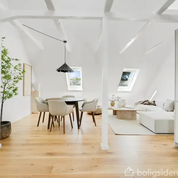 En lys og moderne stue med spisebord og stole i midten, en sofa i højre side, og store vinduer, der giver naturligt lys. En potteplante står ved støttebjælken.