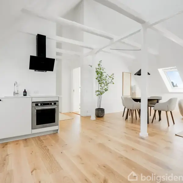 Et lyst, moderne køkken og stueområde med trægulv. Et komfur og sort emhætte i køkkenet. Spisebord med stole i baggrunden. Store ovenlysvinduer giver naturligt lys.
