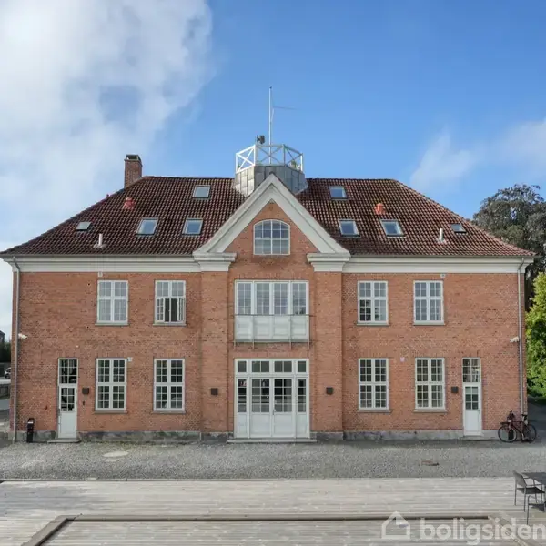 En stor, murstensbygning står stille med rødt tegltag, flere hvide vinduer og en centreret indgangsdør; omgivet af en grusbelagt forplads og grønne træer til højre.