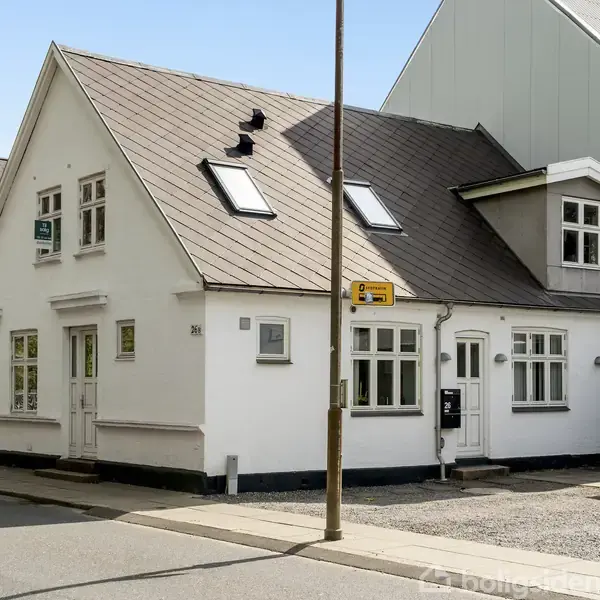 Et hvidt, halvandenetages hus med flere skråvinduer står i et gadehjørne. Et skilt med teksten "Smellof" er placeret på væggen. Omgivet af andre bygninger og fortov.