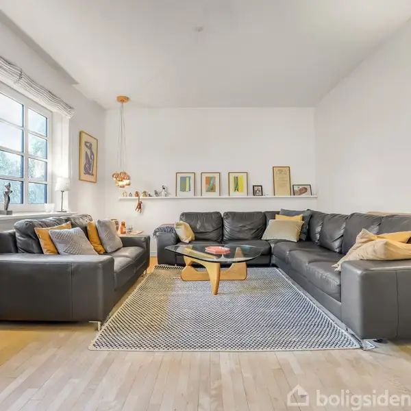 En stue med to store, sorte lædersofaer står i hjørnet omkring et rundt glasbord. Væggene er dekoreret med kunst, og et gulvlampe stråler over tæppet på trægulvet.