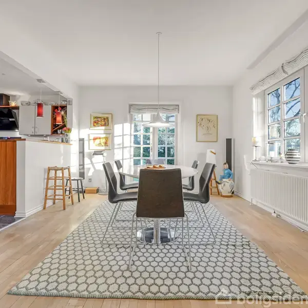 En spisestue med et bord og seks stole, placeret på et tæppe. Rummet er lyst med store vinduer, og der er adgang til et køkken. Lamper og dekorative malerier skaber en hyggelig atmosfære.