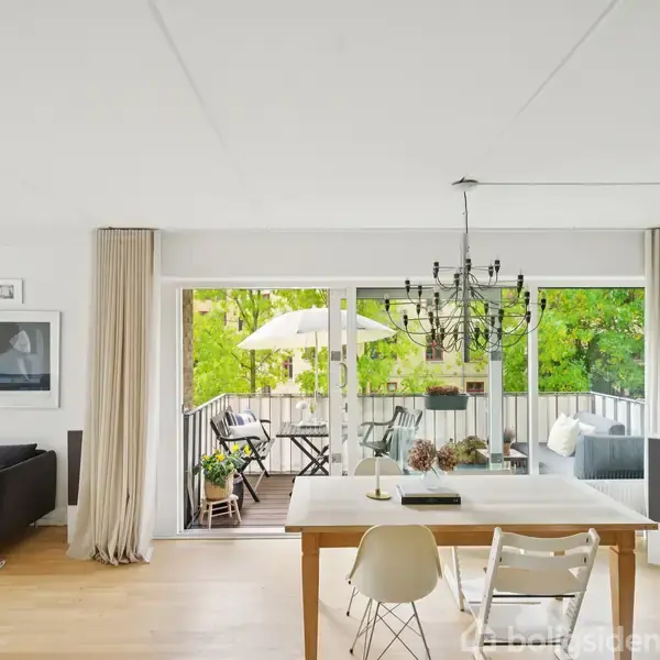Stue med spisebord, omgivet af stole, under en lysekrone. Store glasdøre åbner ud til en altan med parasol og grøn udsigt. Reoler fyldt med bøger til højre.