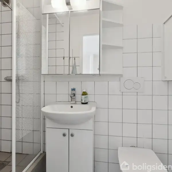 Et moderne badeværelse med en håndvask og skab under, et spejlskab på væggen og en brusekabine i glas. Omgivelserne er hvide fliser.