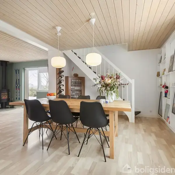 Træbord med sorte stole står i en lys spisestue. En trappe fører opad. Store vinduer og vægkunst præger rummet, der er lyst op af moderne lamper.