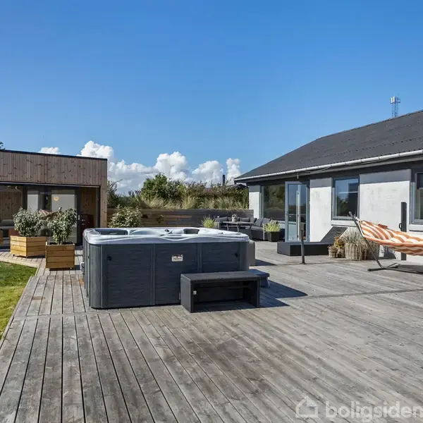 Boblebad og hængekøje på en stor træterrasse ved et moderne hus. I baggrunden ses en lille træbygning og en græsplæne omgivet af grønne træer under en blå himmel.
