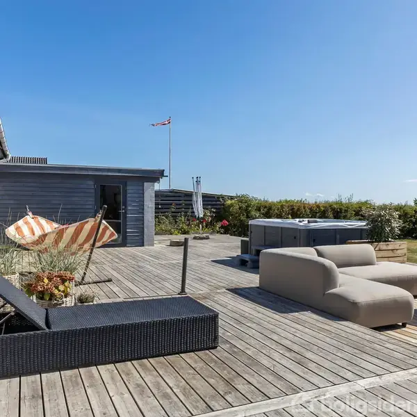 Terrasse med beige sofaer, et rundt bord og en liggestol. En hængekøje med orange striber ses til venstre. Græsplæne og blå himmel i baggrunden.