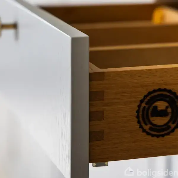 En åben træskuffe med et rundt logo i siden er trukket halvt ud fra et hvidt køkkenskab. Indendørs i et lyst rum.