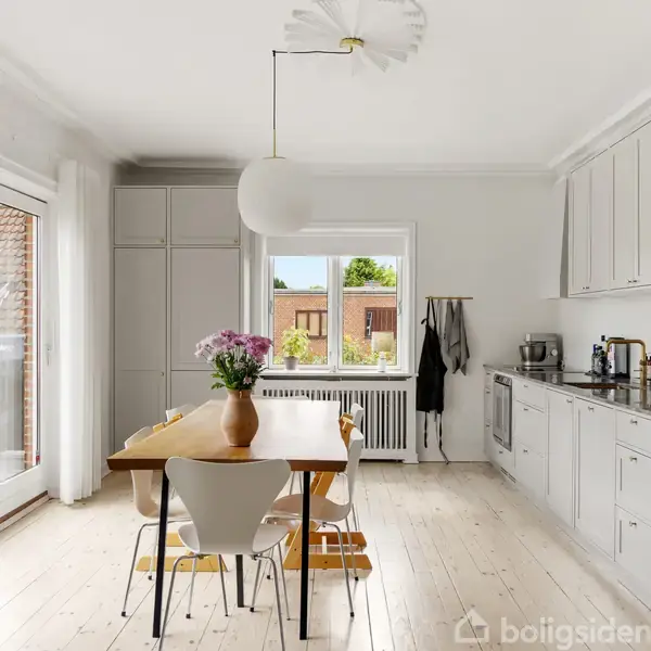 Et moderne køkken med et træbord og hvide stole står i centrum. En vase med blomster står på bordet. Lyse skabe og vinduer med udsigt til haver.