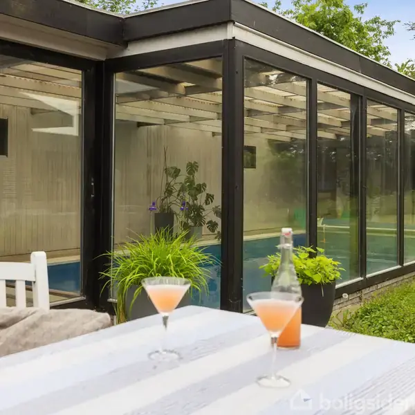To glas med orange drikke står på et bord udenfor ved en glasoverdækket pool. Bordet har en hvid dug, og der er planter omkring. Baggrunden viser træer og natur.