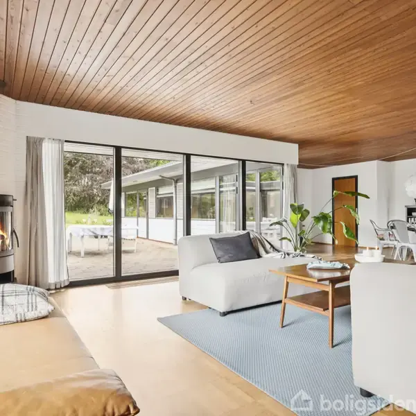 En lys stue med brændeovn til venstre, brændende. Store vinduer viser en terrasse udenfor. Moderne møblering med sofaer og spisebord. Træloft og lyse trægulve skaber en varm atmosfære.