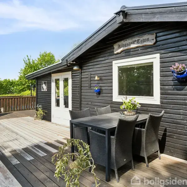 Sort træhus med en terrasse hvor et bord og stole står. En hvid pejs er placeret til venstre. Skiltet "Kingston Cabin" er over vinduet. Grønne planter omkranser terrassen.