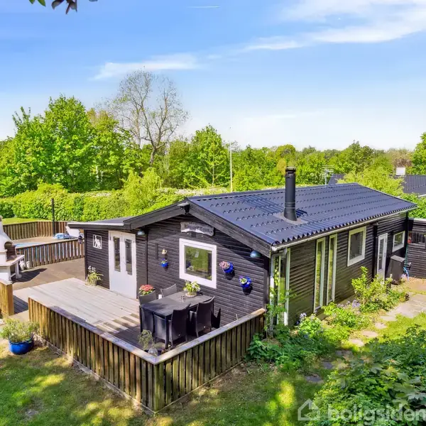 En mørkt træhus med blå vindueskarme, omgivet af grønne træer og buske, står i en græsplæne. Der er en terrasse med borde og stole samt en udendørs grill.