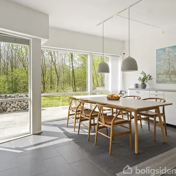 Spisebord med stole er placeret i lys spisestue. Vinduerne åbner op for udsigt til grøn have. Loftslamper hænger over bordet, og maleri pryder væggen.