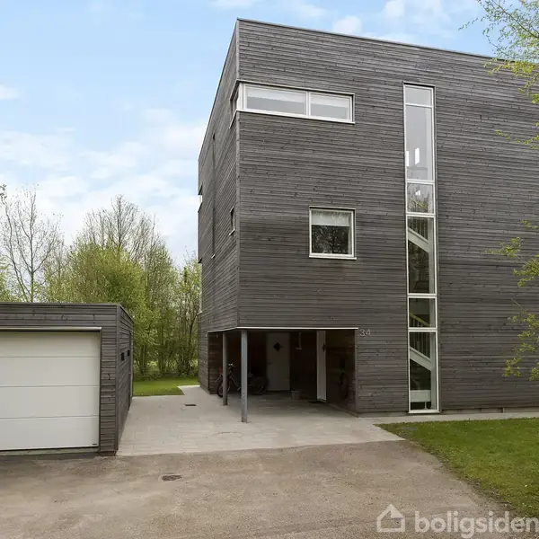 Moderne hus i tre-etager står stille ved siden af en enkeltstående garage. Det er omgivet af træer og grønne områder, hvilket skaber en rolig atmosfære.