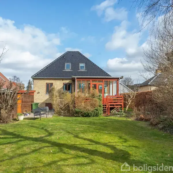 Hus med sort tag og brun murstensfacade. Trappe fører til hævet terrasse med glasvægge. Omgivet af have med græsplæne og buske. Skyfri, klar himmel i baggrunden.