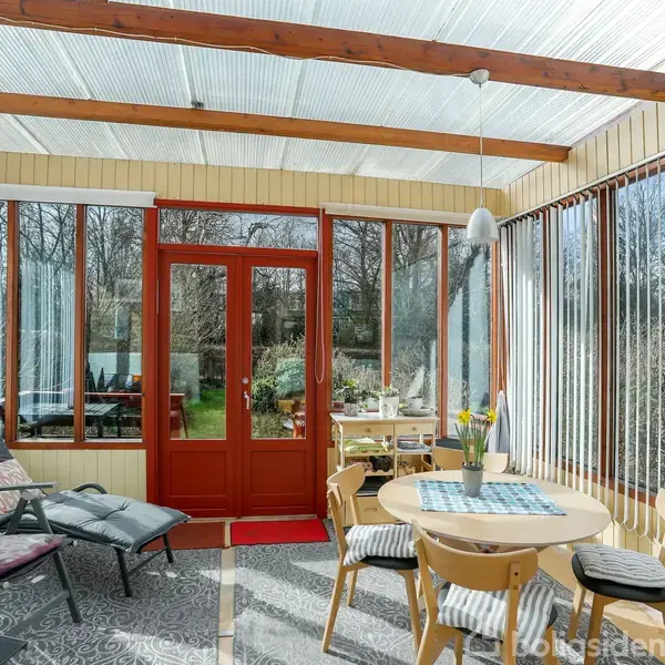 Vinterhave med møbleret bord og stole; blomster i midten. Sollys trænger ind gennem glaslofter og -vægge. Udsigt over have via røde dobbeltdøre. Træpaneler og hyggelig atmosfære.
