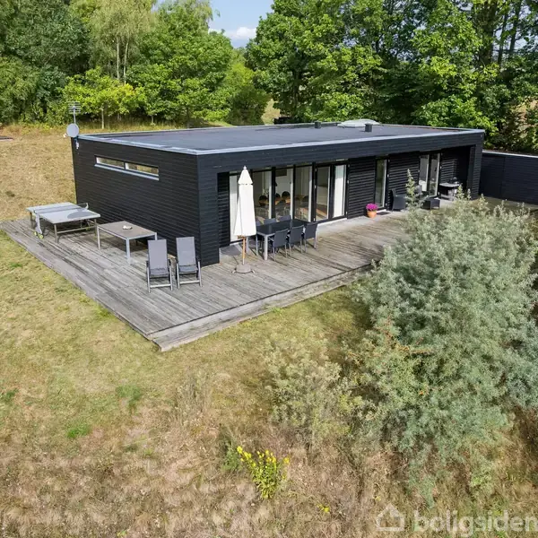 Et sort, rektangulært hus står på en træterrasse med havemøbler og parasol. Det er omgivet af en græsplæne og træer, under en skyfri himmel.